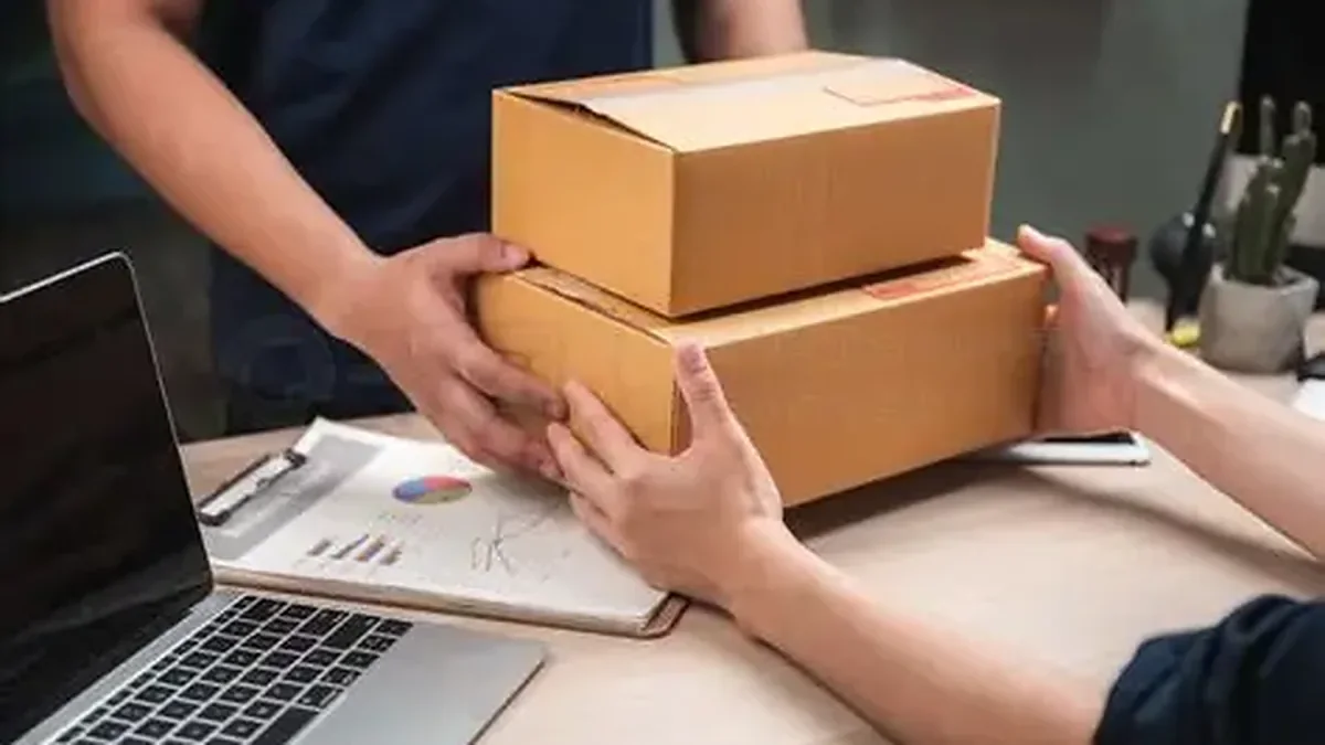 Two people handing cardboard boxes over a desk near a laptop